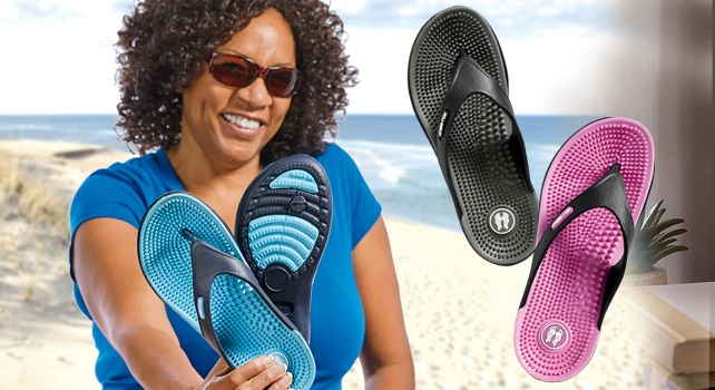 A woman on a beach wearing sunglasses and holding out a pair of blue flip flops. One pink and one black flip flop off to the side.